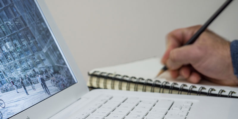 close-up of man writing on notepad next to laptop