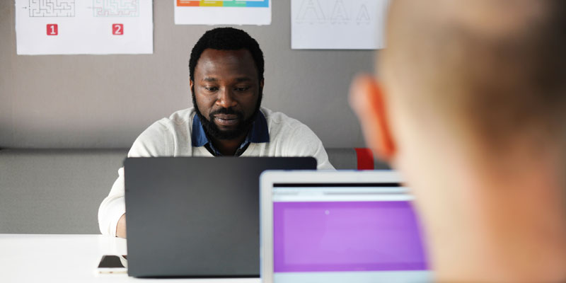 looking over shoulder of two men using laptops sitting in front of each other at conference table