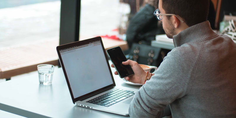 man sitting by window using laptop and phone