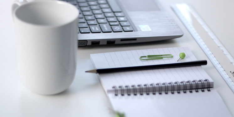 black pencil on ruled notepad beside white ceramic mug and gray laptop computer